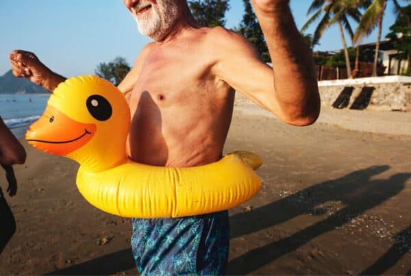 Senior man with gray hair and beard, smiling with a yellow duck floatie at the beach.