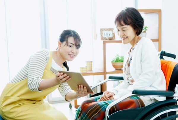 Elderly woman with gray hair smiling in a wheelchair, caregiver with tablet.
