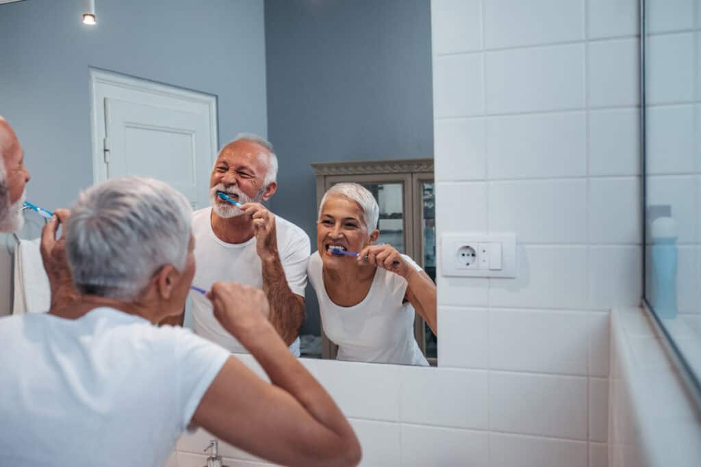 Senior couple brushing teeth in bathroom mirror, embracing aging gracefully.